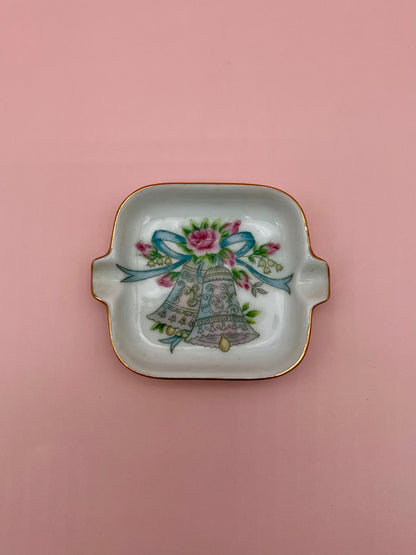 Square white ashtray with gold handles, hand-painted with silver bells tied with a blue ribbon, pink background.