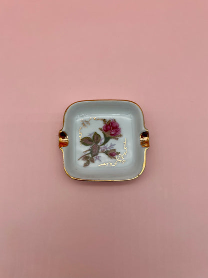 Square white ashtray with gold handles, hand-painted with a single pink rose and gold leaf accents, pink background.