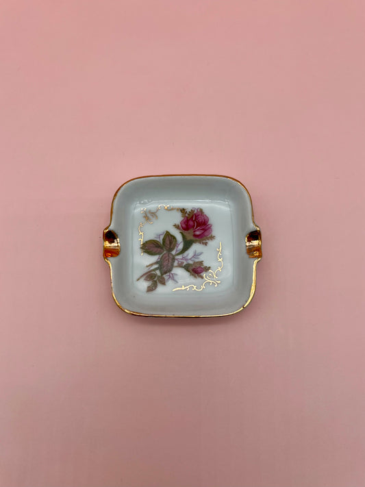 Square white ashtray with gold handles, hand-painted with a single pink rose and gold leaf accents, pink background.