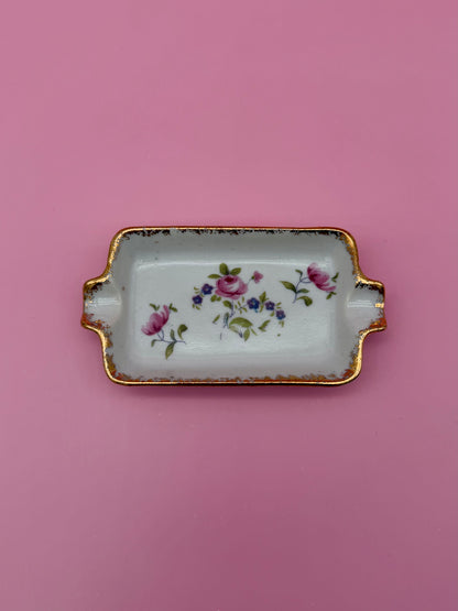 Rectangular white ashtray with gold handles, hand-painted with a small pink and purple floral spray, pink background.