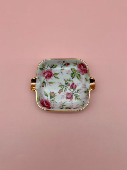 Small square white ceramic ashtray with a pink rose pattern and gold handles, pink background.