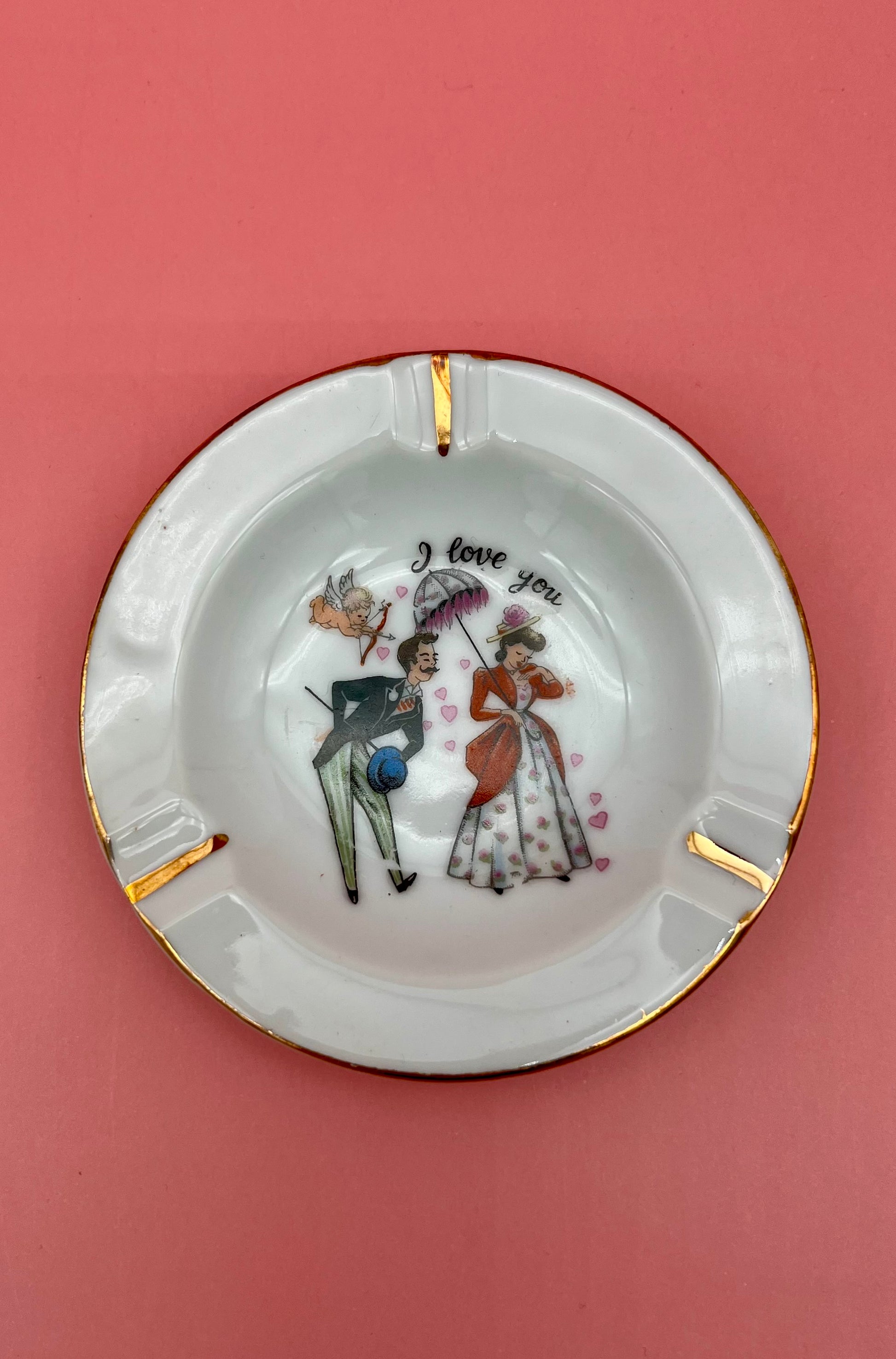 Round white ashtray with gold rim from the Vintage Mini Romance Ashtrays set, hand-painted with a Victorian couple and the caption 'I love you,' pink background.