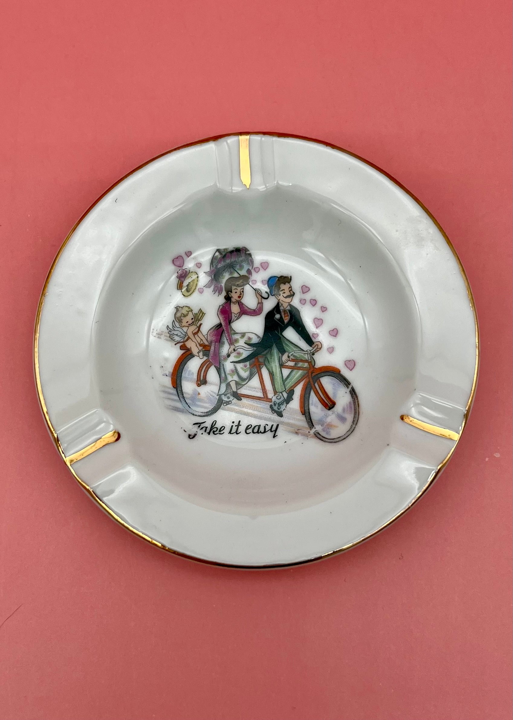 Round white ashtray with gold rim, hand-painted with a couple riding a tandem bicycle and the caption 'Take it easy,' pink background.