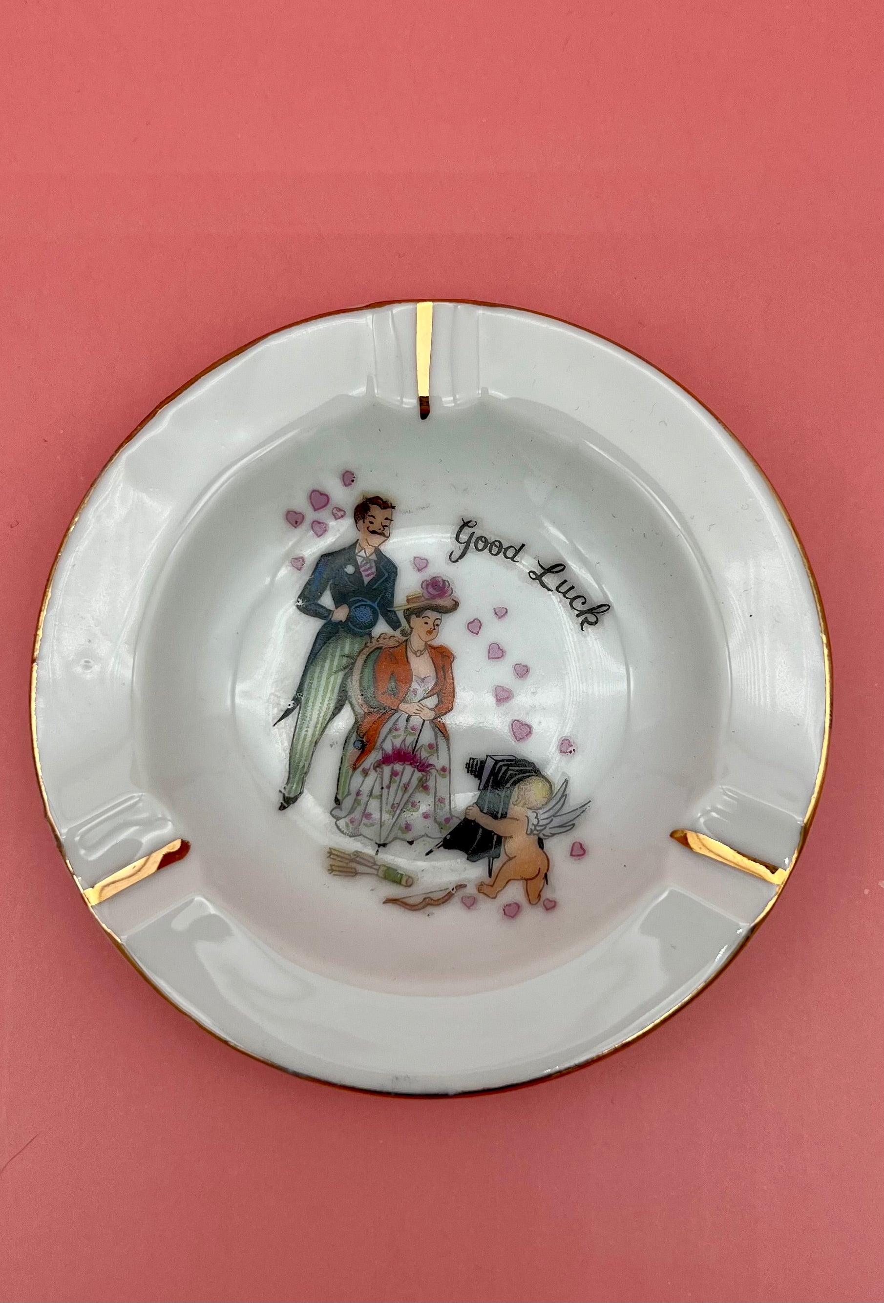 Round white ashtray with gold rim, hand-painted with a couple and a small dog surrounded by hearts, captioned 'Good Luck,' pink background.