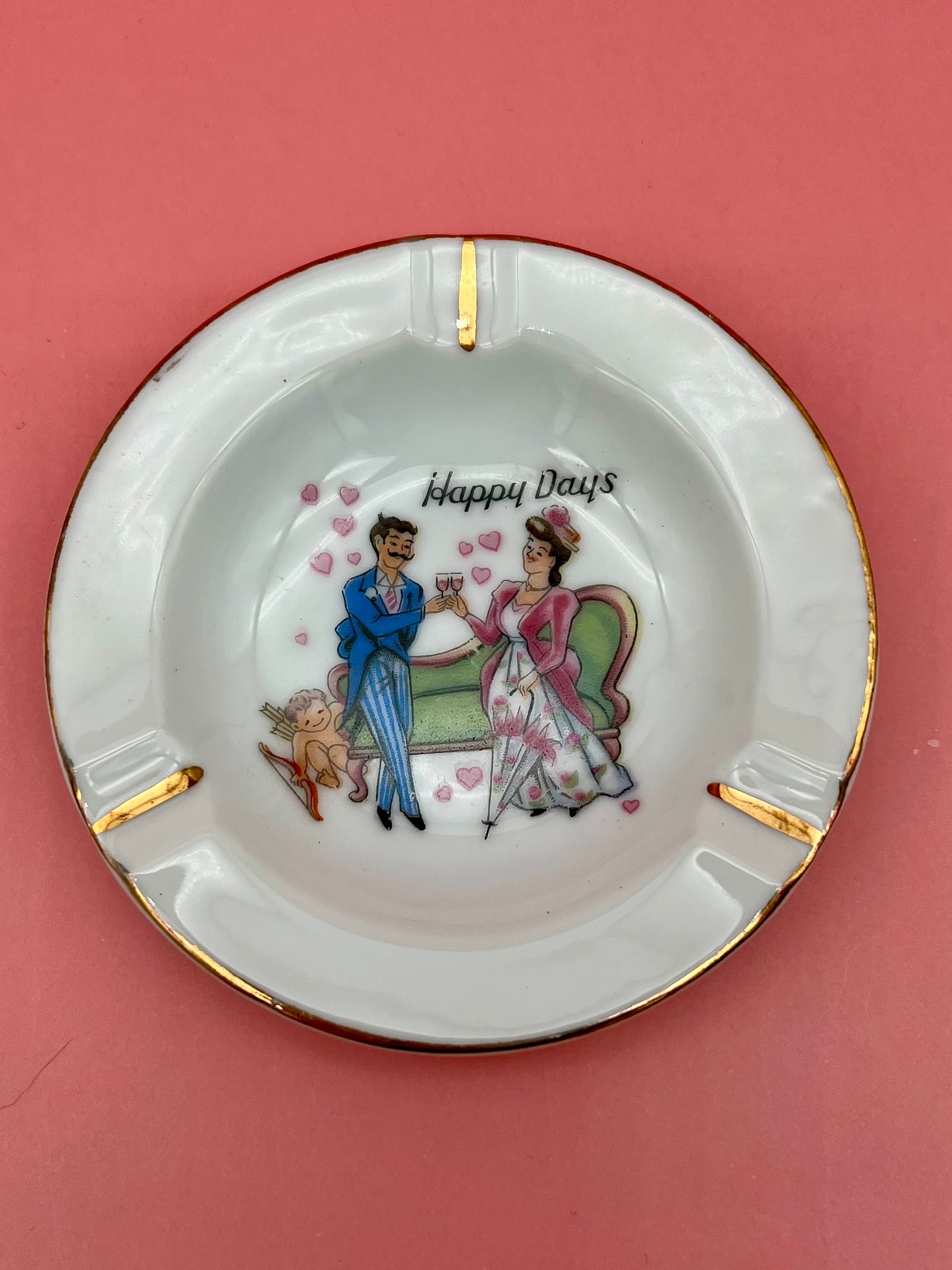 Round white ashtray with gold rim, hand-painted with a couple seated on a bench with a cherub, captioned 'Happy Days,' pink background.