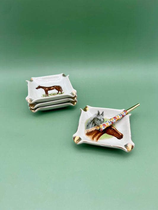 Willa Set of square white ceramic ashtrays each featuring a hand-painted horse illustration, with a glass cigarette pick resting on top, green background.