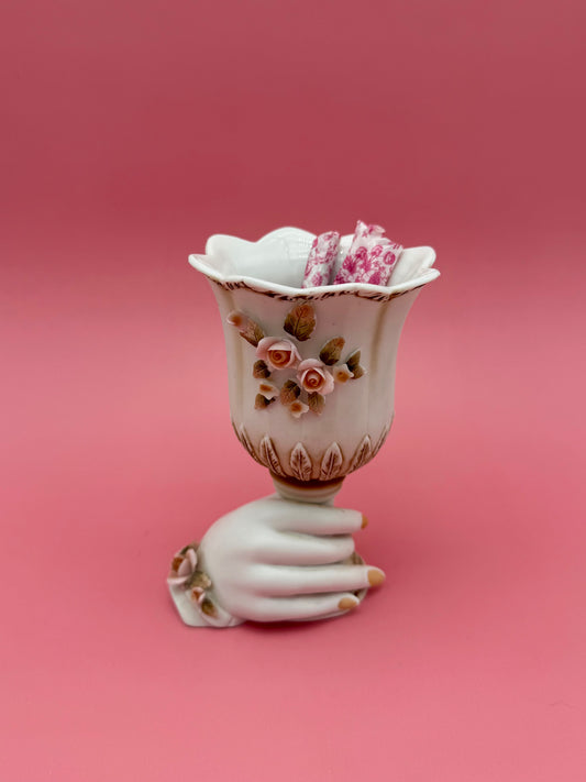 White porcelain ruffled vase resting on a sculpted hand-shaped base, decorated with pink floral accents, dark pink background.